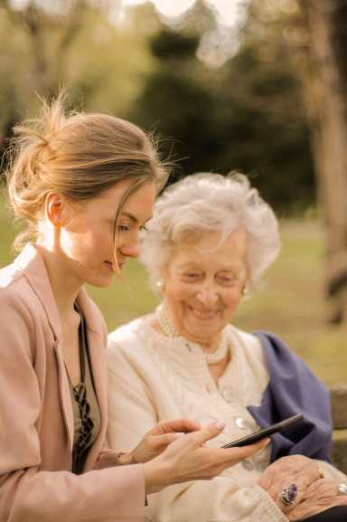 a companion helping an elderly person to use a smartphone, sitting together in a park, illustrating a moment of sharing and kindness outdoors