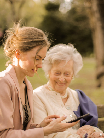 a companion helping an elderly person to use a smartphone, sitting together in a park, illustrating a moment of sharing and kindness outdoors